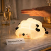 A glowing, squishy sheep-shaped silicone lamp with a calm sleeping face on a marble bedside table.