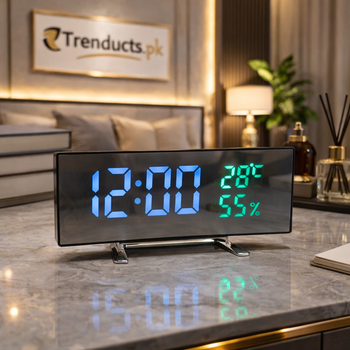 Curved black modern LED digital table clock displaying 12:00 in blue numbers and 28C, 55% weather readouts in green, standing on a wood desk.